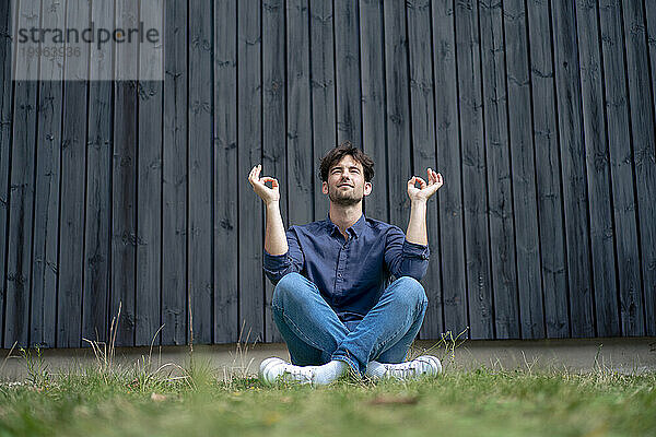Young man meditating sitting on grass in front of wall