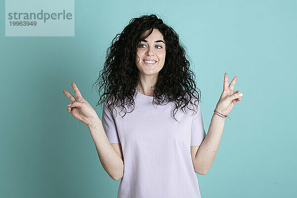 Smiling young woman gesturing peace sign against turquoise background