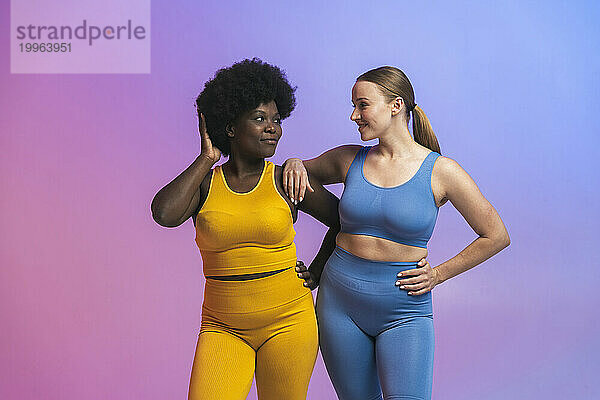 Smiling young multiracial female friends looking at each other while standing against colored background