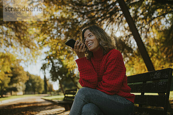 Lächelnde Frau  die im Herbstpark Voicemail auf ihrem Smartphone aufzeichnet