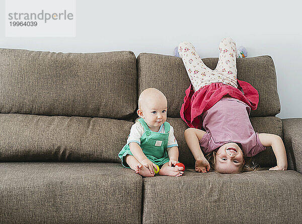 Playful girl doing headstand near baby brother on sofa at home