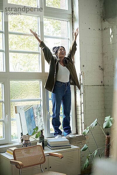 Happy woman with hands raised standing on window sill at home