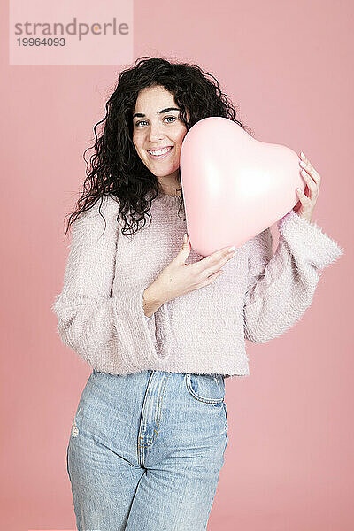 Glückliche schöne Frau  die mit einem herzförmigen Ballon vor rosa Hintergrund steht