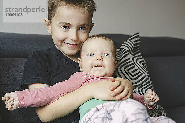 Smiling boy holding baby sister on sofa at home