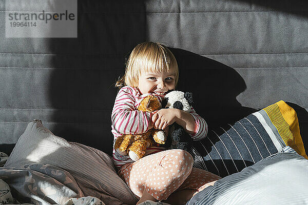 Cheerful girl hugging stuffed toys on bed at home