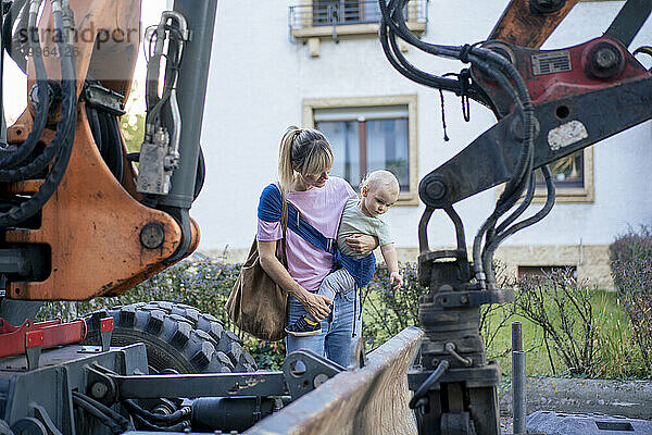 Mutter trägt Sohn  der neben Bulldozer steht