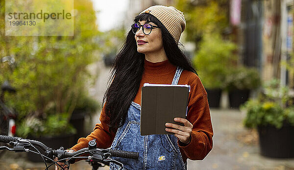 Lächelnde Frau hält Tablet-PC in der Hand und steht mit Fahrrad