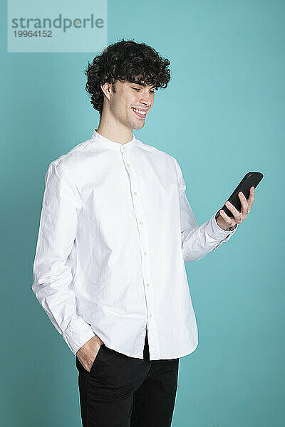 Lächelnder Mann mit der Hand in der Tasche und Smartphone vor cyanfarbenem Hintergrund