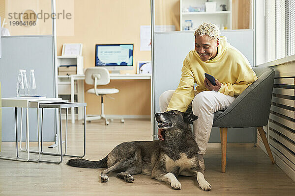 Lächelnde Frau hält Smartphone und streichelt Hund in Tierklinik