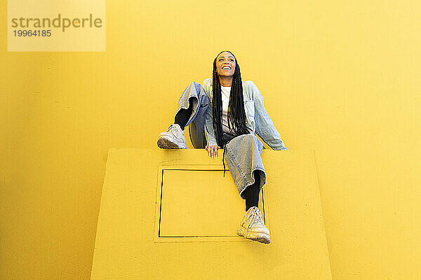 Glückliche junge Frau sitzt in der Nähe der gelben Wand