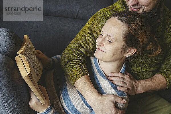 Lächelnde Frau liegt auf dem Schoß eines Mannes und liest zu Hause ein Buch