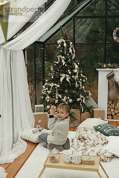 Fröhlicher Junge  der zu Hause auf dem Teppich neben dem Weihnachtsbaum spielt