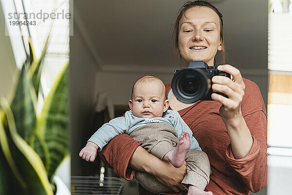 Spiegelbild einer lächelnden Mutter  die ihren Sohn trägt und mit der Kamera fotografiert