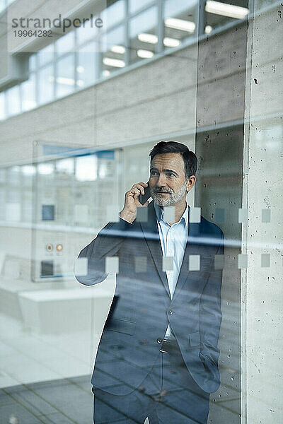 Lächelnder reifer Geschäftsmann  der durch Glas auf dem Mobiltelefon spricht