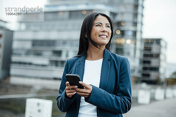 Lächelnde Geschäftsfrau steht mit Mobiltelefon vor dem Gebäude