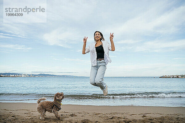 Verspielte Frau gestikuliert Friedenszeichen und springt neben Pudelhund am Strand