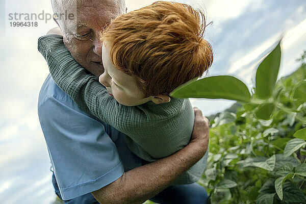 Liebevoller Großvater umarmt und spielt mit Enkel in der Nähe von Pflanzen auf dem Feld