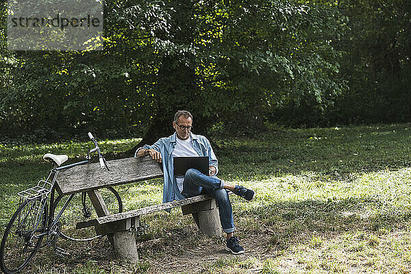 Mann sitzt mit Laptop auf Bank im Park