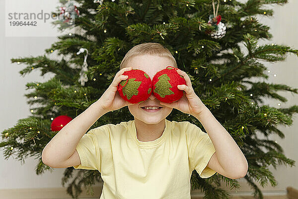 Lächelnder Junge versteckt Augen mit Weihnachtsschmuck zu Hause