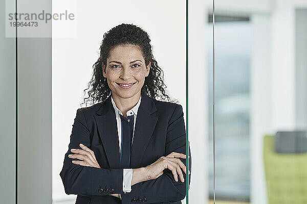 Happy businesswoman standing with arms crossed near glass door in office