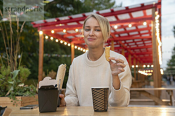 Lächelnde blonde Frau isst Essen am Tisch im Vergnügungspark