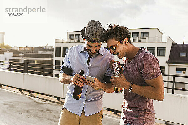 Lächelnde Männer benutzen Smartphone und halten Bierflaschen auf dem Dach
