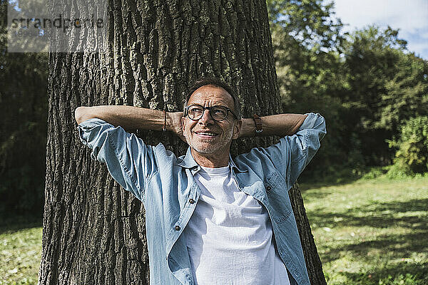 Nachdenklicher Mann mit Händen hinter dem Kopf vor Baum im Park