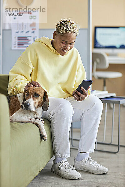 Lächelnde Frau benutzt Smartphone und streichelt Hund auf Sofa in Tierklinik