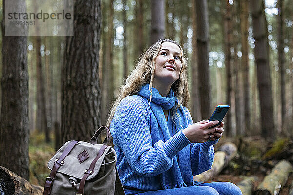 Lächelnde Frau sitzt mit Rucksack und Smartphone im Cannock Chase Forest