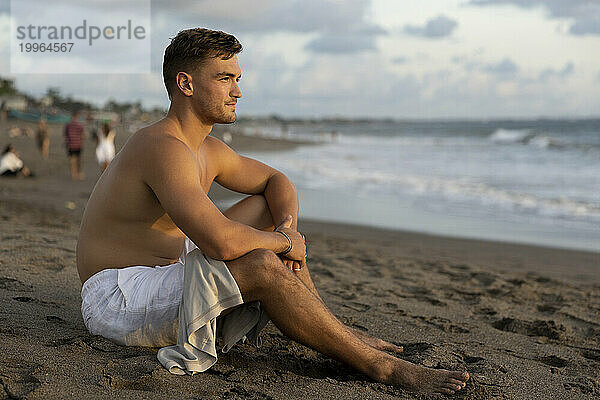 Shirtless young man sitting on sand at beach