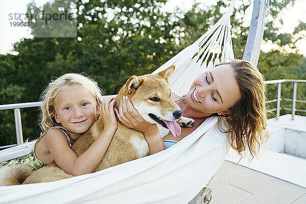 Lächelnde Mutter und Tochter streicheln den Hund Shiba Inu in der Hängematte