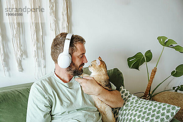 Mann mit Katze trägt Kopfhörer und sitzt zu Hause auf der Couch