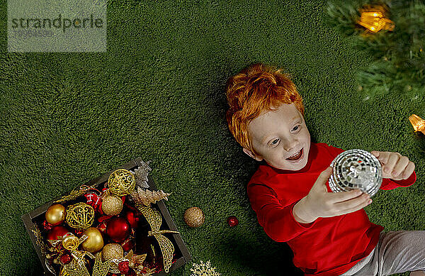 Glücklicher rothaariger Junge  der mit Weihnachtskugel auf grünem Teppich spielt