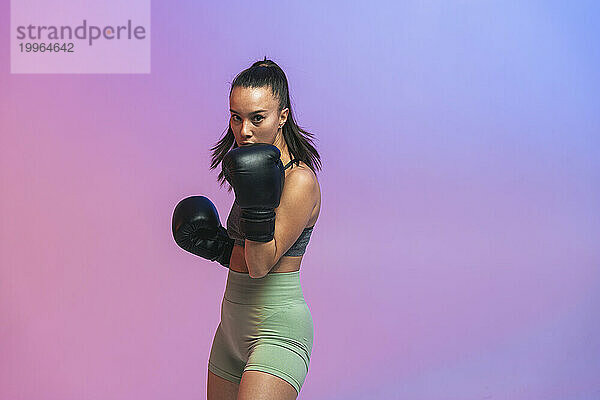 Selbstbewusste junge Frau trainiert mit schwarzen Boxhandschuhen vor farbigem Hintergrund