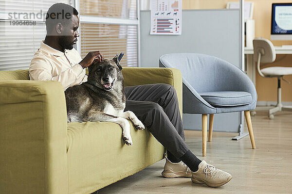 Junger Mann benutzt Mobiltelefon und sitzt mit Hund auf dem Sofa in der Klinik