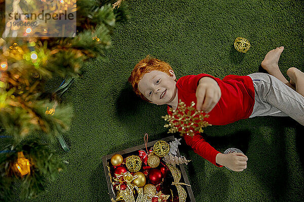 Glücklicher rothaariger Junge  der mit Weihnachtsschmuck auf grünem Teppich spielt