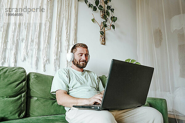 Mann mit Kopfhörern sitzt zu Hause auf der Couch und benutzt Laptop