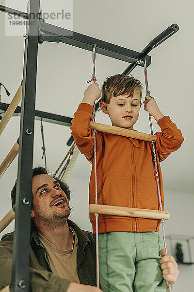 Smiling father teaching son to climb rope ladder at home