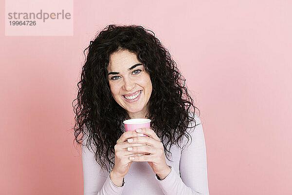 Glückliche Frau hält Einwegkaffeetasse vor rosa Hintergrund