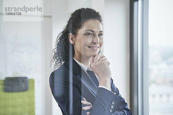 Smiling mature businesswoman with hand on chin seen through glass
