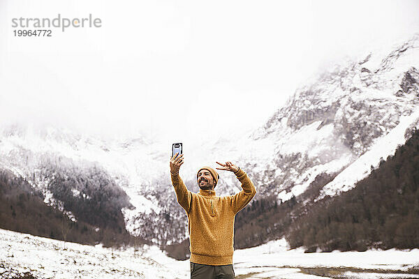 Lächelnder Mann zeigt ein Friedenszeichen und macht ein Selfie mit seinem Smartphone vor den Pyrenäen  Katalonien  Spanien