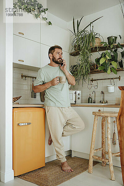 Mann mit Kaffeetasse telefoniert zu Hause in der Küche