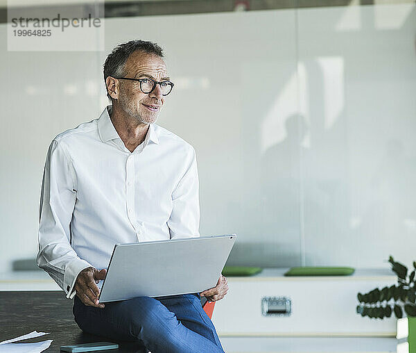 Lächelnder älterer Geschäftsmann sitzt mit Laptop auf dem Schreibtisch