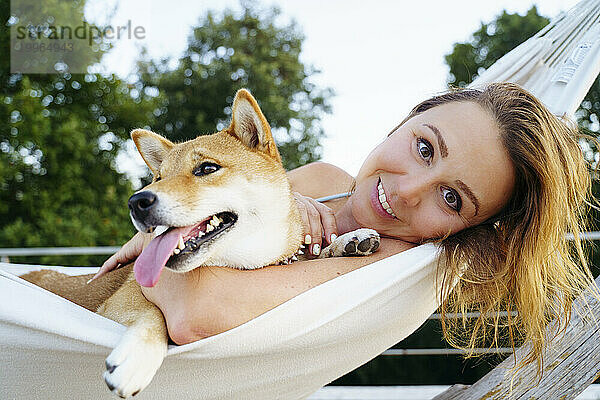 Lächelnde blonde Frau liegt mit Shiba Inu Hund in der Hängematte