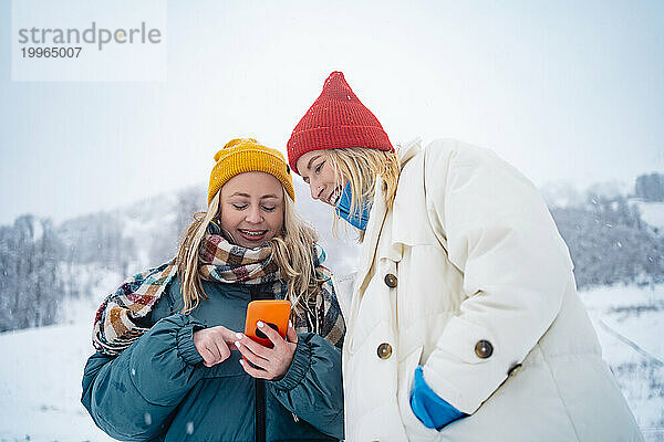 Lächelnde Frau  die ihr Smartphone auf einem schneebedeckten Berg benutzt und einem Freund zeigt