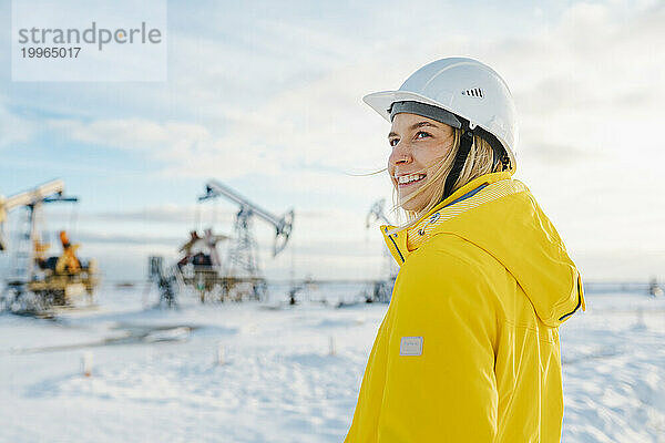 Lächelnder junger Ingenieur mit Schutzhelm auf dem Ölförderfeld im Winter