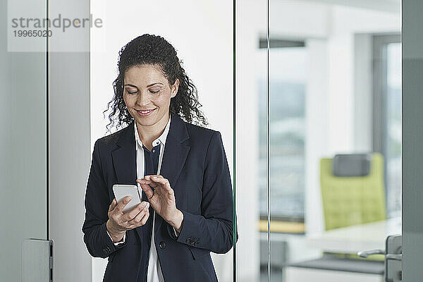 Lächelnde reife Geschäftsfrau benutzt Smartphone im Büroeingang