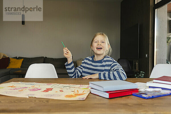 Glückliches Mädchen hält Bleistift in der Hand und sitzt mit einer Zeichnung am Schreibtisch