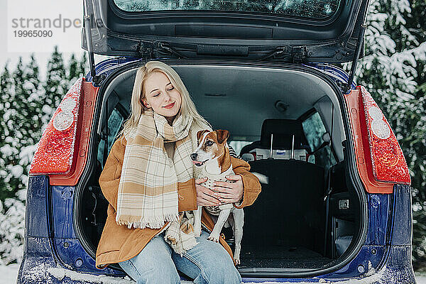 Lächelnde Frau sitzt mit Hund im Kofferraum eines Autos