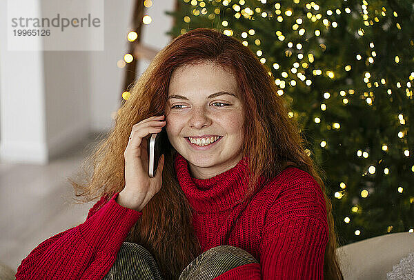 Lächelnde Frau telefoniert zu Hause neben dem Weihnachtsbaum